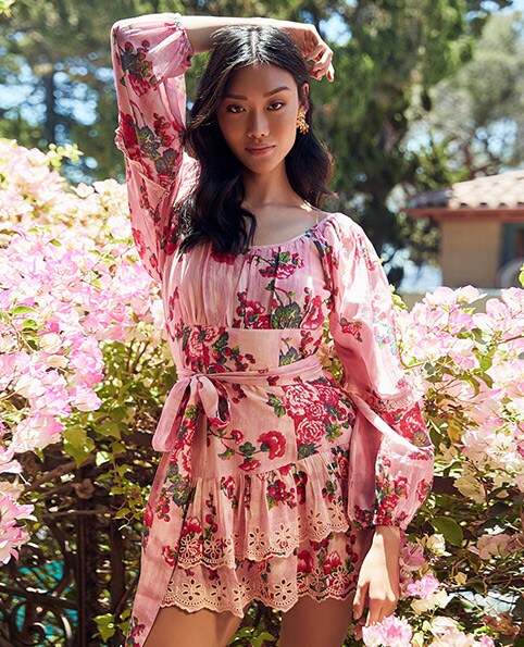 A model standing in front of flowers with one arm over her head wearing a pink long sleeve mini dress with a floral print, waist tie and ruffle hem. Pretty Dress Roundup. Shop the Edit.