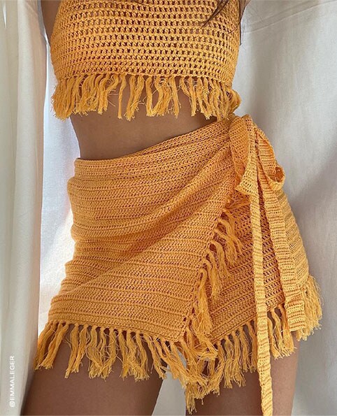 A close-up photo of Emma Leger wearing a yellow crochet crop top with fringe trim and a matching mini skirt with wrap styling. Matching Sets. Shop Now.
