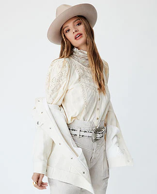 A model wearing a beige hat, a cream button up blouse, a white denim jacket, beige pants with a white studded belt. The New Western. Shop Now.